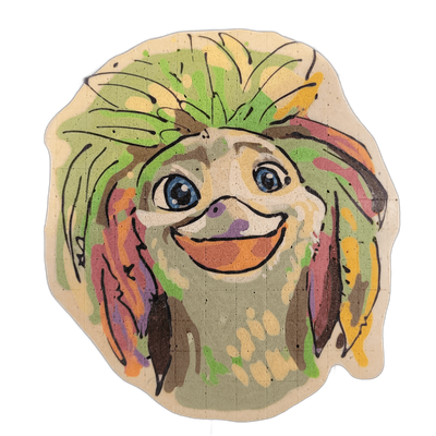 Pancake art of Javan, a cartoon-style creature face with large blue eyes, a wide open-mouthed grin, a small rounded nose, and long shaggy fur or hair radiating outward in all directions. Drawn by Dan with bold black outlines and dense fill in green, yellow, orange, brown, purple, and muted tan, wit…