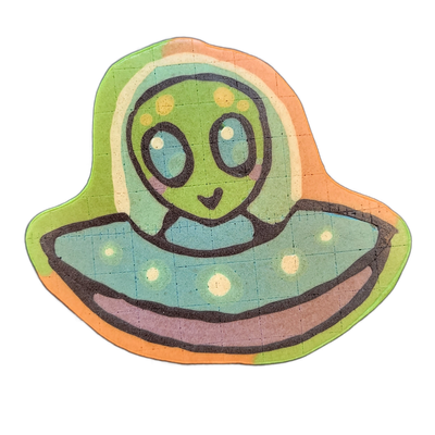 Pancake art depicting Trav, a JoP original character — a green-skinned alien with large dark oval eyes, small dot markings on the forehead, and a small curved mouth, seated inside the dome of a flying saucer with a wide flat brim and a rounded lower hull.
