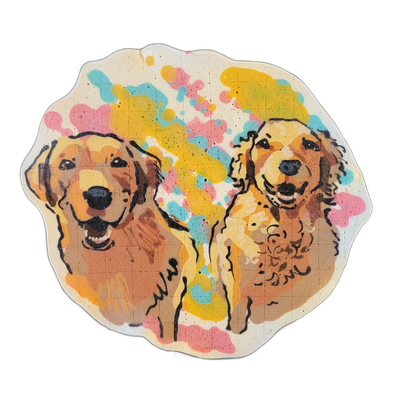 Pancake art depicting two golden-colored dogs, Rory and Zeus, shown side by side from roughly the chest up, each facing forward with open mouths; the dog on the left has straighter fur and the one on the right has curlier fur. Bold black outlines define the forms, filled with warm amber and brown t…