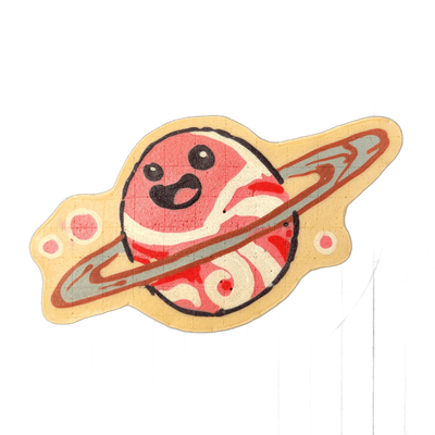 Saturnina, a JoP original character depicted as a cartoon planet with Saturn-style rings and a smiling face with large black eyes and an open mouth, shown with two small circular shapes flanking the planet body against a pale yellow background shape. The pancake art uses bold black outlines, a pale…