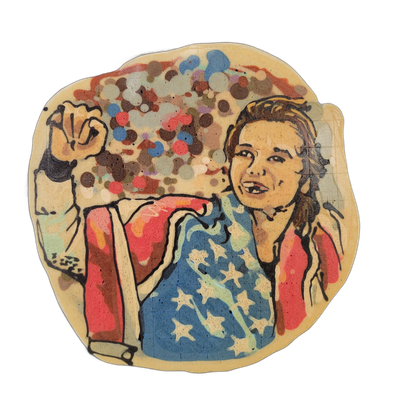 Pancake art depicting a young woman with brown hair in a braid, wearing a blue top with white stars and a red jacket, raising her right fist in the air against a background of multicolored confetti dots in red, blue, brown, teal, and white. Rendered by Dan with bold dark outlines and broad color fi…