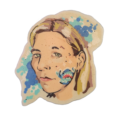 Pancake art portrait of a person named La, showing a forward-facing face with green eyes, light skin, freckles, blonde hair pulled back, and an abstract blue and teal creature or face painted on the lower left cheek area, with blue and teal paint-splash-style batter shapes extending around the head…
