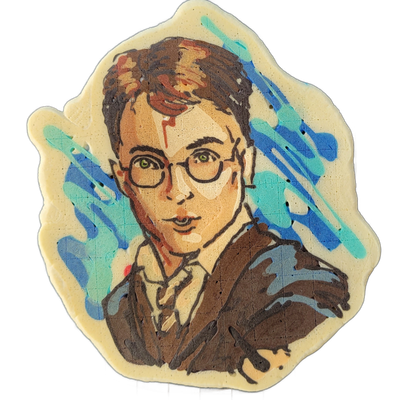 A pancake art portrait of a young male figure with short dark brown hair, round wire-frame glasses, bright green eyes, a lightning-bolt scar on the forehead, and a dark brown jacket with a light collared shirt and tie visible at the neck; blue and teal brushstroke-style shapes fill the background b…
