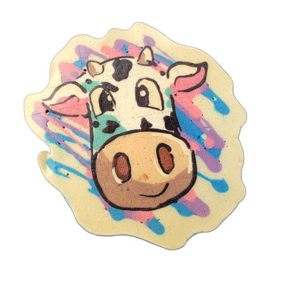 A cartoon cow face centered on a scallop-edged pancake, facing forward, with large round eyes, black-and-white spotted markings on the forehead and top of the head, small horns, pink inner ears, and a large rounded brown muzzle with two oval nostrils and a slight upward curve at the mouth. The piec…