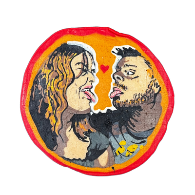 Two people face each other with mouths open and tongues out in a playful expression, a small red heart drawn between them at the top center; the figure on the left has long wavy brown and golden hair and bare shoulders, while the figure on the right has short dark hair, a full beard, and a tattooed…