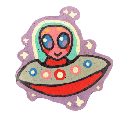 Pancake art depicting Trav, a JoP original character: a round-headed pink alien with large blue oval eyes, small dot markings on the forehead, and a slight smile, seated in a gray and red flying saucer decorated with circular blue and pink porthole shapes, set against a purple background scattered…