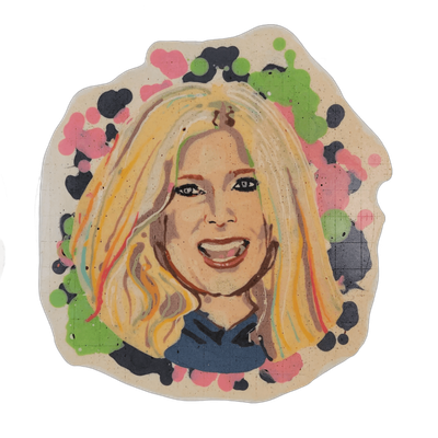 Pancake art portrait of a woman with long, wavy blonde hair, dark eye makeup, and an open-mouthed smile, wearing what appears to be a dark blue top, centered within an irregular cream-colored border filled with clusters of green, pink, and dark navy splatters and blobs. Dan rendered the face with f…
