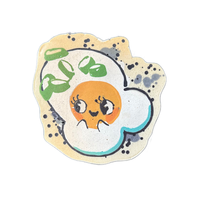 Pancake art of Sunny, a JoP original character depicted as a kawaii-style fried egg with an orange yolk face featuring large lashed eyes, rosy cheeks, and a small smile, topped with a crown of green onion slices rendered in layered green tones, set against a white egg-white body with a teal accent…