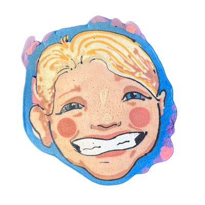 Pancake art of a smiling child's face named Sarah, with short blonde hair, blue eyes, rosy circular cheeks, freckles, and a wide open-mouth smile showing white teeth. Wella rendered the piece with bold black outlines, peach and orange skin tones, yellow and orange hair strokes, and a blue irregular…