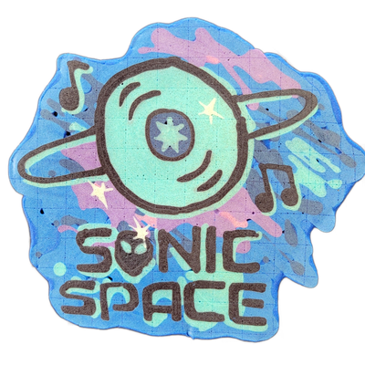 Pancake art depicting a circular vinyl record styled as a planet with a ring orbiting it, flanked by musical notes and small stars, with the text 'SONIC SPACE' lettered below in bold rounded characters. Rendered by Dan with bold dark outlines on a mint green and teal fill, set against a background…