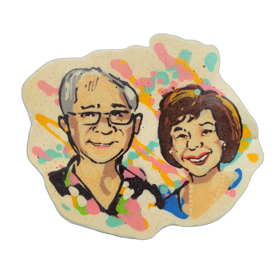 Pancake art depicting two adults shown from the shoulders up, side by side: on the left, a man with short gray hair and dark-framed rectangular glasses wearing a dark collared shirt; on the right, a woman with short brown hair wearing a blue top and a small earring, shown smiling with her mouth ope…
