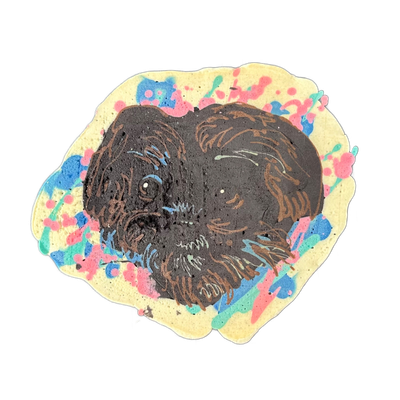 A pancake art portrait of a dark-coated dog's face, rendered in dark brown batter on a cream-colored oval base, surrounded by splatters and blobs of pink, blue, and teal batter. Fine brown lines define the fur texture, ears, and facial features including a visible eye with a white highlight; bold o…