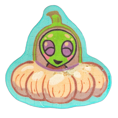 Pancake art depicting Trav, a JoP original character — a cartoon alien figure with a round green head, large purple oval eyes, rosy cheeks, and a small smile, wearing a purple scarf and emerging from the top of a wide, ribbed pumpkin-shaped flying saucer rendered in peach and brown tones, with a gr…