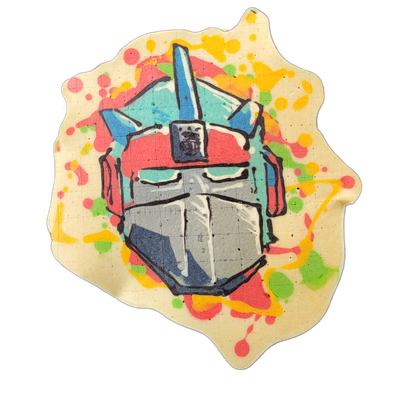 A pancake art piece depicting a robotic humanoid face with angular mechanical features, including a flat faceplate with a centered vertical ridge, teal rectangular eye lenses, a dark rectangular panel on the forehead, and blue fin-like helmet crests extending upward and to the sides, with red ear-s…