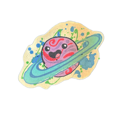 Saturnina (Nina), an original character from the JoP cast depicted as a cartoon planet with a smiling face and Saturn-style rings encircling her body, illustrated in pancake art by Wella on a flat-cut beige batter base with paint-splatter-style batter accents in blue and green surrounding the compo…