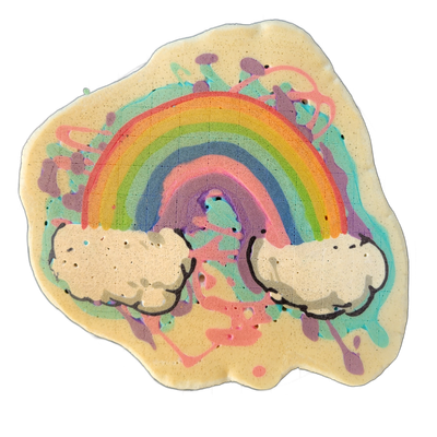 A pancake art piece depicting a rainbow arching between two white fluffy clouds, rendered on a roughly circular, uneven pancake base with a pale yellow cook color. The rainbow is illustrated with bold curved bands in red, orange, yellow, green, blue, and violet; the clouds are outlined in dark batt…