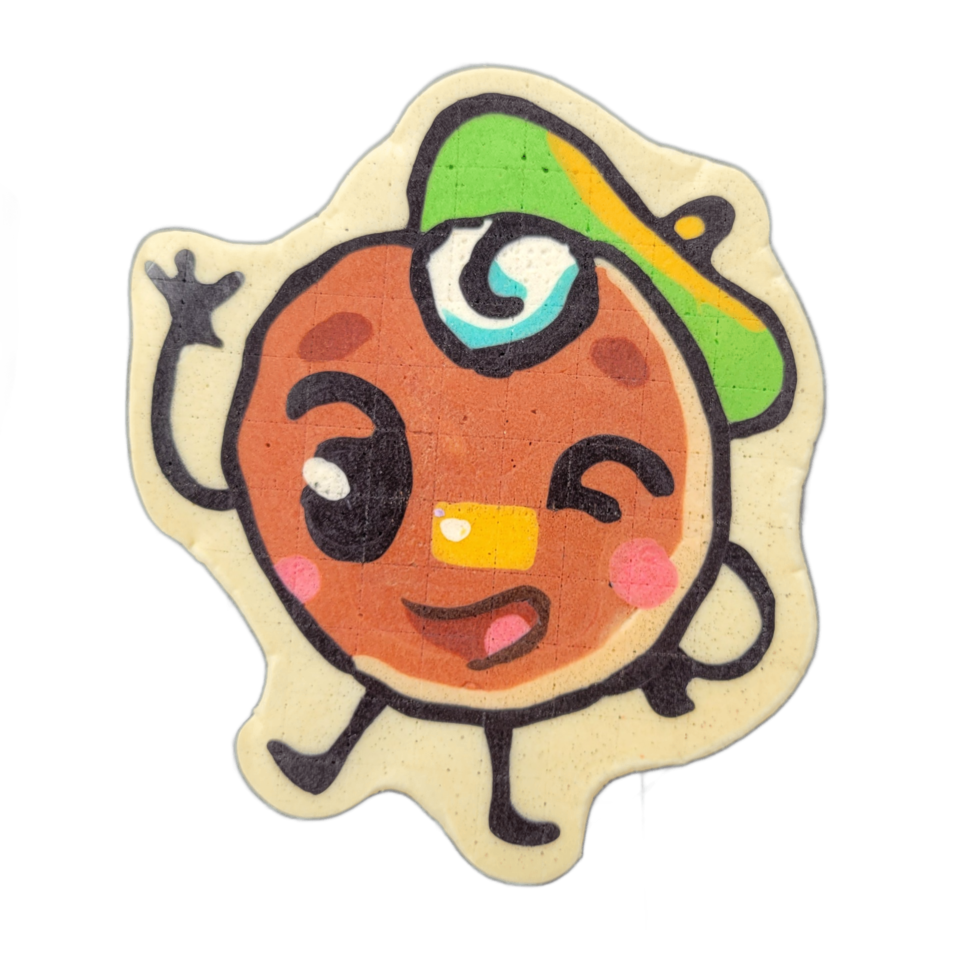 Pancake art depicting Jacques Floppes, a round cartoon character with a brown circular face, one large open eye with a teal spiral iris and one closed eye in a wink, rosy cheek spots, an open mouth showing a pink interior and a yellow tooth, a green and yellow cap worn with the brim tilted to the s…