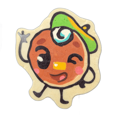 Pancake art depicting Jacques Floppes, a round cartoon character with a brown circular face, one large open eye with a teal spiral iris and one closed eye in a wink, rosy cheek spots, an open mouth showing a pink interior and a yellow tooth, a green and yellow cap worn with the brim tilted to the s…