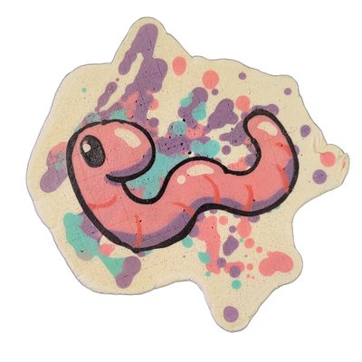 Pancake art depicting Hermie the Wurmy, a pink worm character with a round head, one visible eye with a white highlight, and a body that curves in an S-shape with short curved line details along its length, set against a white background scattered with irregular paint-splatter shapes in violet, cor…