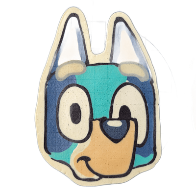Pancake art depicting the head of Bluey, a cartoon blue heeler puppy character, facing forward with two pointed ears, large round white eyes with dark pupils, one eye appearing half-closed, a tan muzzle, and a small dark nose. Rendered by Dan using bold dark outlines and flat color fills in shades…