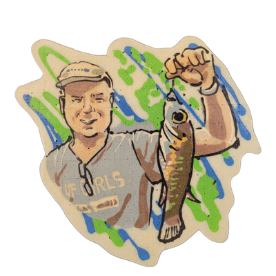 Pancake art depicting a smiling adult man wearing a cap and a gray short-sleeve shirt with text reading 'OF GIRLS' or similar lettering, holding up a fish by its tail with his raised right fist, the fish rendered with a visible eye, yellow and brown coloring, and spine detail. Bold black outlines d…