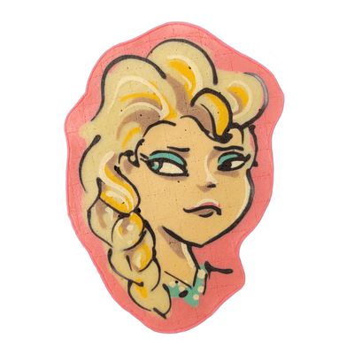Pancake art depicting a cartoon female face with voluminous blonde hair styled in a side braid, teal eye shadow, and a small teal snowflake detail near the collar, set against a coral-pink background shaped loosely around the figure. Bold black outlines define the hair, facial features, and braid,…