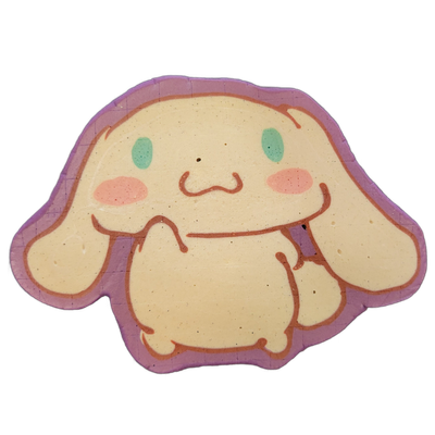 Pancake art depicting a round-bodied cartoon character with very long drooping ears, teal oval eyes, pink oval cheek markings, a small curved nose, and stubby limbs held up in front of the body, set against a violet border that follows the character's silhouette. The piece uses bold reddish-brown o…