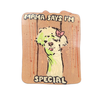 Pancake art depicting a llama facing forward with fluffy white and light green batter forming its body and face, a pink bow on top of its head, and a slightly open mouth; the piece is framed with vertical dark lines suggesting a fence or gate, and block text reading 'MAMA SAYS I'M' across the top a…