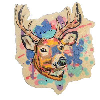 A deer head with antlers, facing slightly left, rendered as a cut-out piece of pancake art resting on a dark gridded craft mat. Bold black outlines define the deer's face, ears, and antlers, filled with warm browns, oranges, and yellows; the background area surrounding the head is splattered with l…