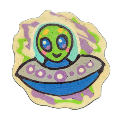 Trav, a green-skinned alien character with large blue eyes, pink cheek marks, and yellow spots on the top of the head, sits visible from the torso up inside a gray flying saucer decorated with purple circle details and a blue underside, set against a cream-colored scalloped background with loose pa…