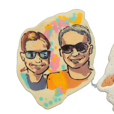 Pancake art depicting two people wearing sunglasses posed close together, the figure on the left with short reddish-brown hair and blue-mirrored sunglasses, the figure on the right with short gray hair and dark sunglasses, both showing slight smiles; the left figure wears a gray-blue top and the ri…