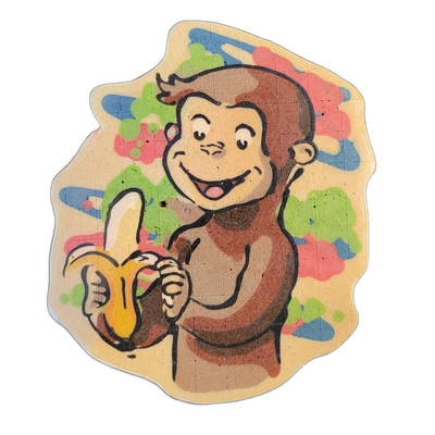 Pancake art depicting Curious George, the cartoon monkey, shown from the chest up, holding a partially peeled banana in one hand and smiling with an open mouth; the figure has large round ears, a tuft of hair on top of the head, and a lighter-toned face and chest contrasting with darker brown fur o…