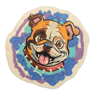 A circular pancake art piece depicting a bulldog's face in a cartoon style, shown from the front with an open mouth, tongue out, and a spiked red collar visible at the bottom of the neck. The piece uses bold black outlines filled with orange, brown, white, and pink batter for the dog's face, set ag…