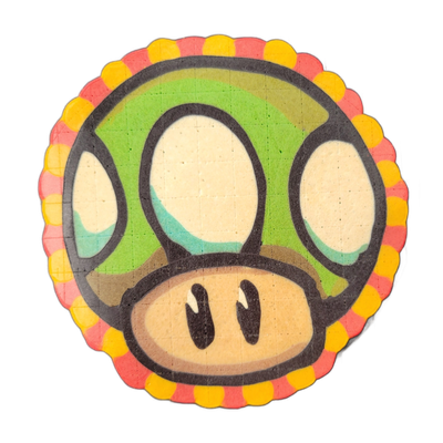 A circular pancake art piece depicting a cartoon mushroom character face with a large domed green cap featuring white oval spots, a beige rounded face with two dark nostril-like marks, and a wide beige lower section; the border of the circle is decorated with alternating red and yellow scalloped do…