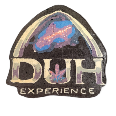Pancake art of a shield-shaped badge design featuring the large text 'DUH' in the center and 'EXPERIENCE' in smaller letters beneath it, with an arch of white lines framing the upper portion and an abstract cloud-like form rendered in blue, violet, and pink above a small cannabis leaf motif between…