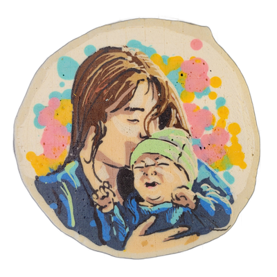Pancake art depicting a person with long brown hair leaning their face down toward a newborn baby wearing a green knit hat; the person holds the baby against their chest with both hands, and the baby's mouth is open with fists raised near its face. The figures are rendered with bold black outlines…