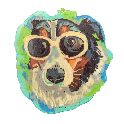 Pancake art depicting a dog's face wearing large round-lensed glasses with a white frame, shown from the front, with fluffy dark fur on top and around the sides, tan and rust-colored markings on the cheeks and ears, and a white muzzle area. The piece is rendered with bold outlines and flat color fi…