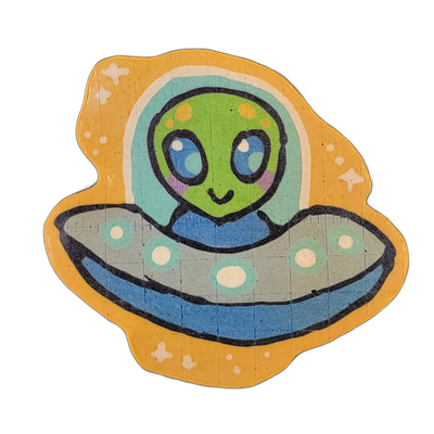 Pancake art of Trav, a green-skinned alien character with large blue eyes, rosy cheeks, and a small smile, seated inside a gray flying saucer with teal and white circular markings and a blue underside; the saucer's transparent dome is rendered in light teal, and Trav wears a blue scarf visible at t…