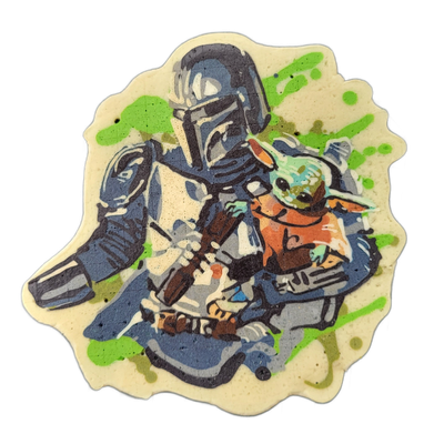 Pancake art depicting two figures: a tall armored figure in a blue-gray helmet and beskar plate armor holding a small green-faced creature with large ears wearing an orange robe, based on the subject identified as Mando and Grogu. The art uses bold dark outlines with color fills in blue-gray, brown…