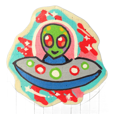 Pancake art depicting Trav, a green cartoon alien with large dark oval eyes, rosy cheek markings, and a small smile, shown from the torso up inside a gray flying saucer with alternating red and green circular lights along its rim and a blue underside band; a pink oval halo frames the figure behind…