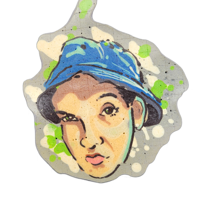Pancake art depicting a young woman's face wearing a blue bucket hat, with dark hair visible at the sides, brown eyes, and slightly parted lips with pink coloring; the face is surrounded by an irregular gray border with scattered green and white paint-like splatters. The piece uses bold black outli…
