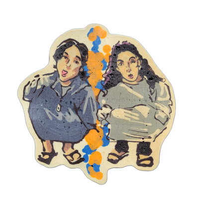 Pancake art depicting two young girls with dark hair seated side by side, both with open mouths and wide eyes, wearing casual tops and sandals; a vertical splash of orange and blue batter runs between them, with purple curls visible in the hair of the figure on the right. The piece uses bold dark o…