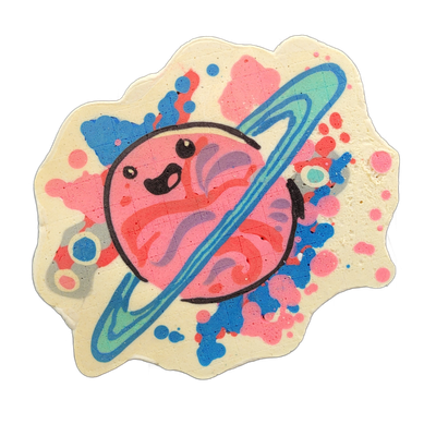 Saturnina, a JoP original character depicted as a round pink planet with a cartoon face — two small dot eyes and an open curved mouth — encircled by a tilted ring rendered in blue and teal, set against a cream background scattered with splatters and dots in pink, red, blue, and lavender. The pancak…