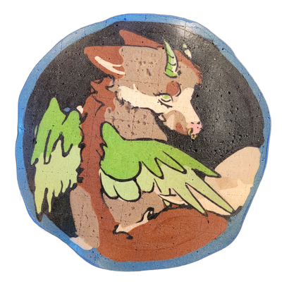 Pancake art depicting Dehli, a winged and horned maned wolf character used as a social media mascot, shown in profile view with a brown and tan fur body, green feathered wings spread to either side, a small green horn, green eyes, a pink nose, and a gold ear accent against a dark background with a…