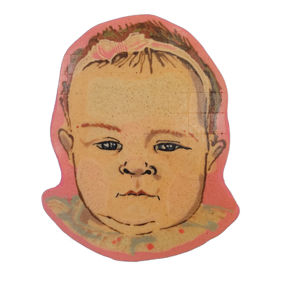 Pancake art portrait of Judgy Margot, an original character depicted as a baby's face with sparse hair, a small bow headband, blue-gray eyes, and a neutral closed-mouth expression, wearing a ruffled collar with small dot details at the base of the frame. Dan rendered the piece with fine dark brown…