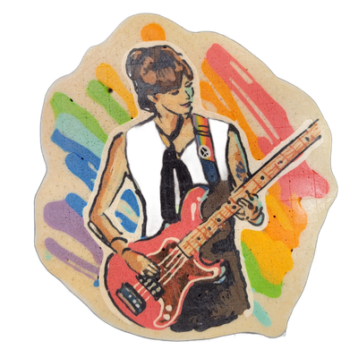 Pancake art depicting a figure named Nikko, shown from roughly the waist up, holding a red electric guitar with a maple-colored neck; the figure has short brown hair, wears a dark sleeveless top with a small circular pin on the chest, a dark neck tie or scarf, and has visible tattoos on one arm, wi…