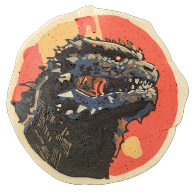 Pancake art depicting Godzilla, shown as a close-up portrait of a large reptilian creature's head and upper body in profile, mouth open wide with visible teeth and tongue, a single red eye visible, and a row of dorsal spines along the top of the head and back. The image uses bold black outlines fil…
