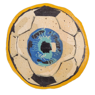 Pancake art depicting a soccer ball with a large stylized eyeball at its center, surrounded by a yellow circular border; the soccer ball is rendered in cream and dark brown-black with bold black outlines defining the panel shapes, and the central eye features a blue and teal iris with dark swirling…
