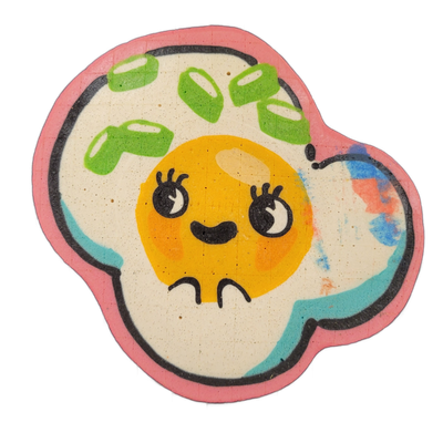 Pancake art depicting Sunny, an original JoP character designed as a fried egg with a face: a round orange yolk forms the head, drawn with large cartoon eyes featuring prominent lashes, rosy circular cheeks, and a small curved mouth, with short curved lines below suggesting a chin or neck; the yolk…