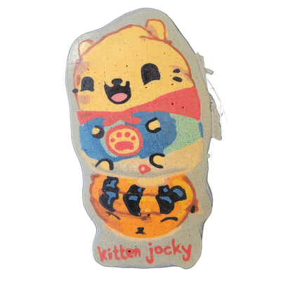 Pancake art of a cartoon cat character labeled 'Kitten Jocky' in red lettering along the bottom; the cat has a round yellow head with black dot eyes, an open mouth with a pink tongue, small ears, and wears a red bow tie, a blue patterned vest or jacket with a pink paw-print emblem on the chest, and…